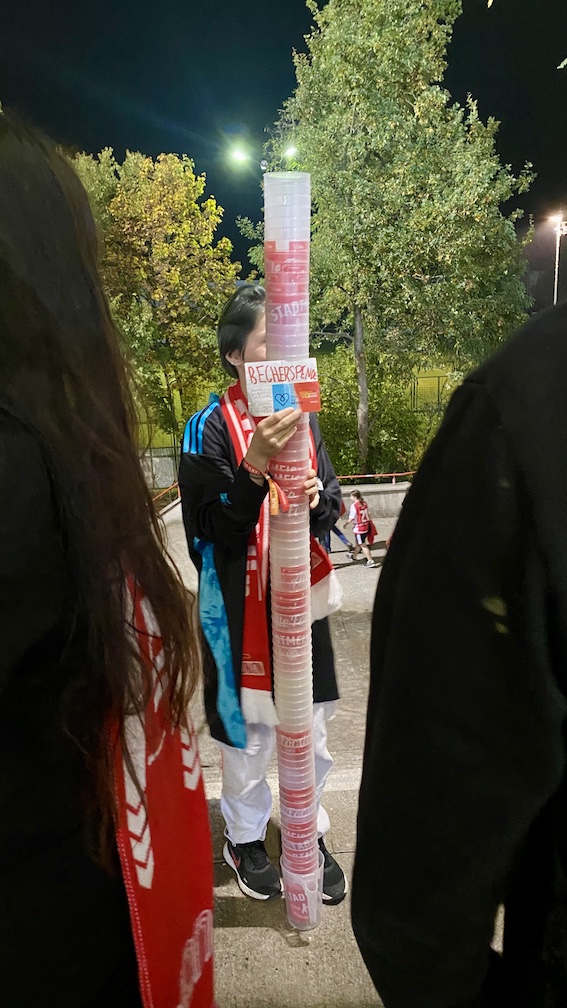 Foto vom Sammeln nach einem Flutlichtspiel am oberen Ende der Treppe von Sektor 3. In der Mitte ist ein Turm aus Trinkbechern zu sehen, der schon deutlich über den Kopf der sammelnden Jungunioerin hinausragt. Außerdem der weiter unten im Text beschriebene, eher kleine Zettel.