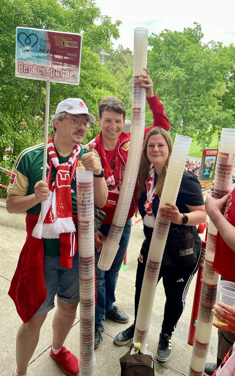 Foto einiger lieber Freund*innen, die uns an diesem Spieltag beim Sammeln vertreten haben. Sie stehen am oberen Ende der westlichen Treppe von Sektor 3 und halten 4 große Bechertürme, die sehr viele Spenden für die Stiftung bedeutet haben.